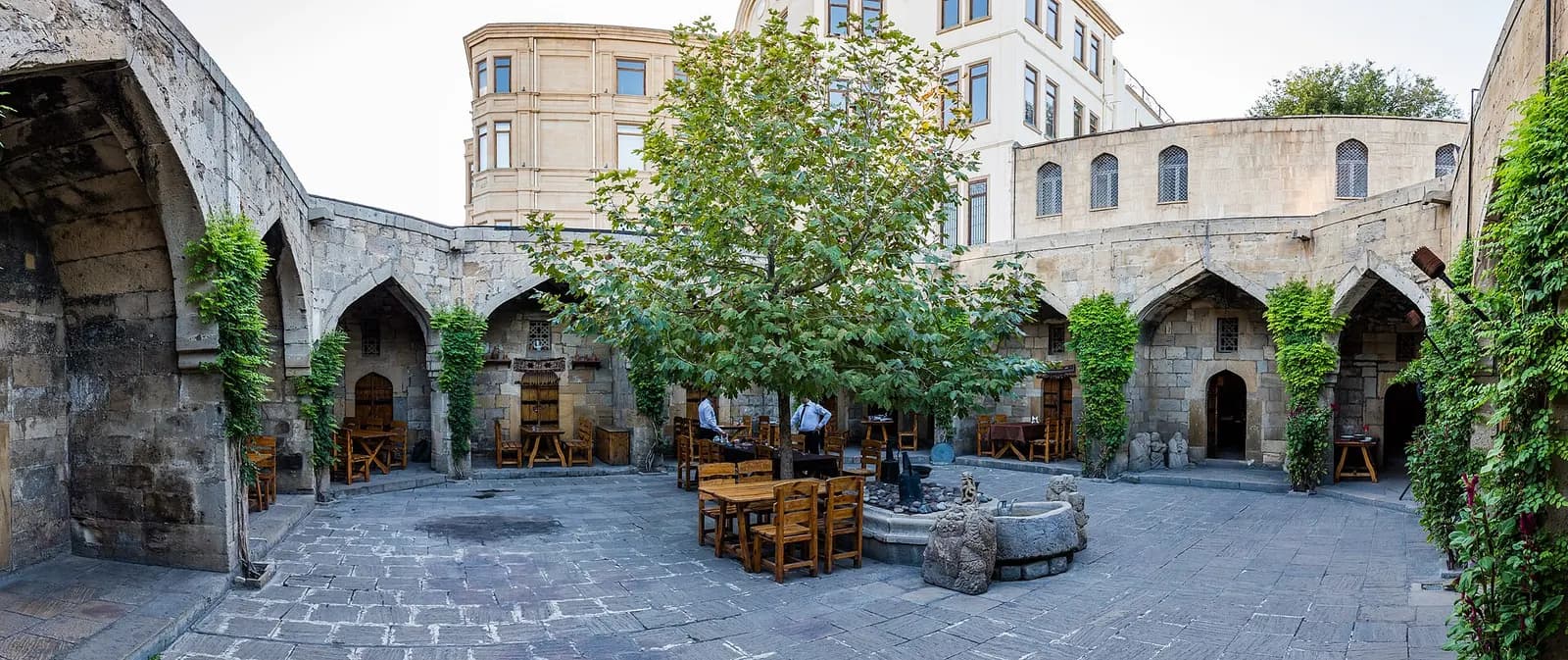 Stone arcaded courtyard of a caravanserai in Baku's Old City, with tables under a tree and climbing plants on the arches