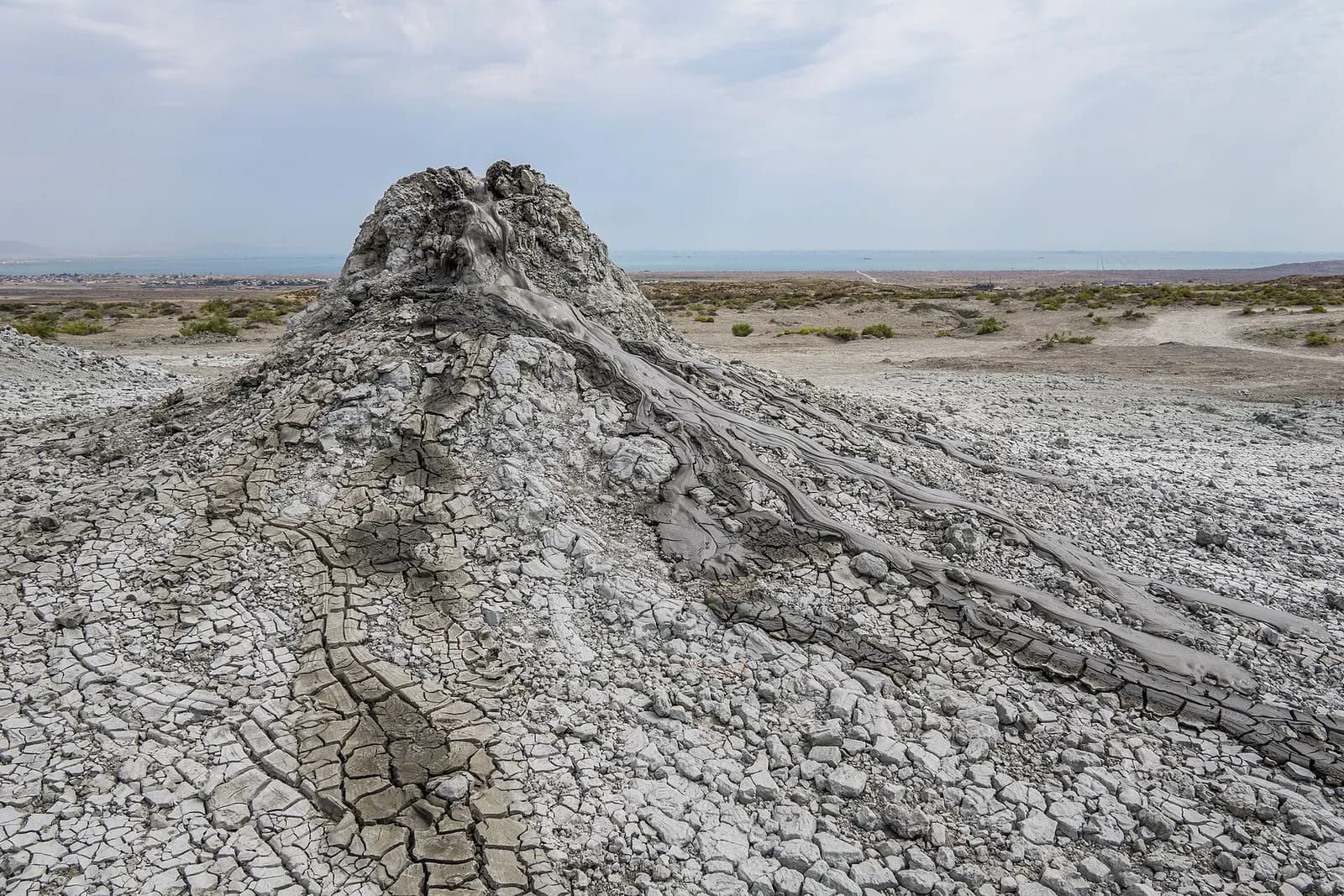 A grey mud volcano cone at Gobustan with fresh mud flowing down its cracked sides and the Caspian Sea on the horizon