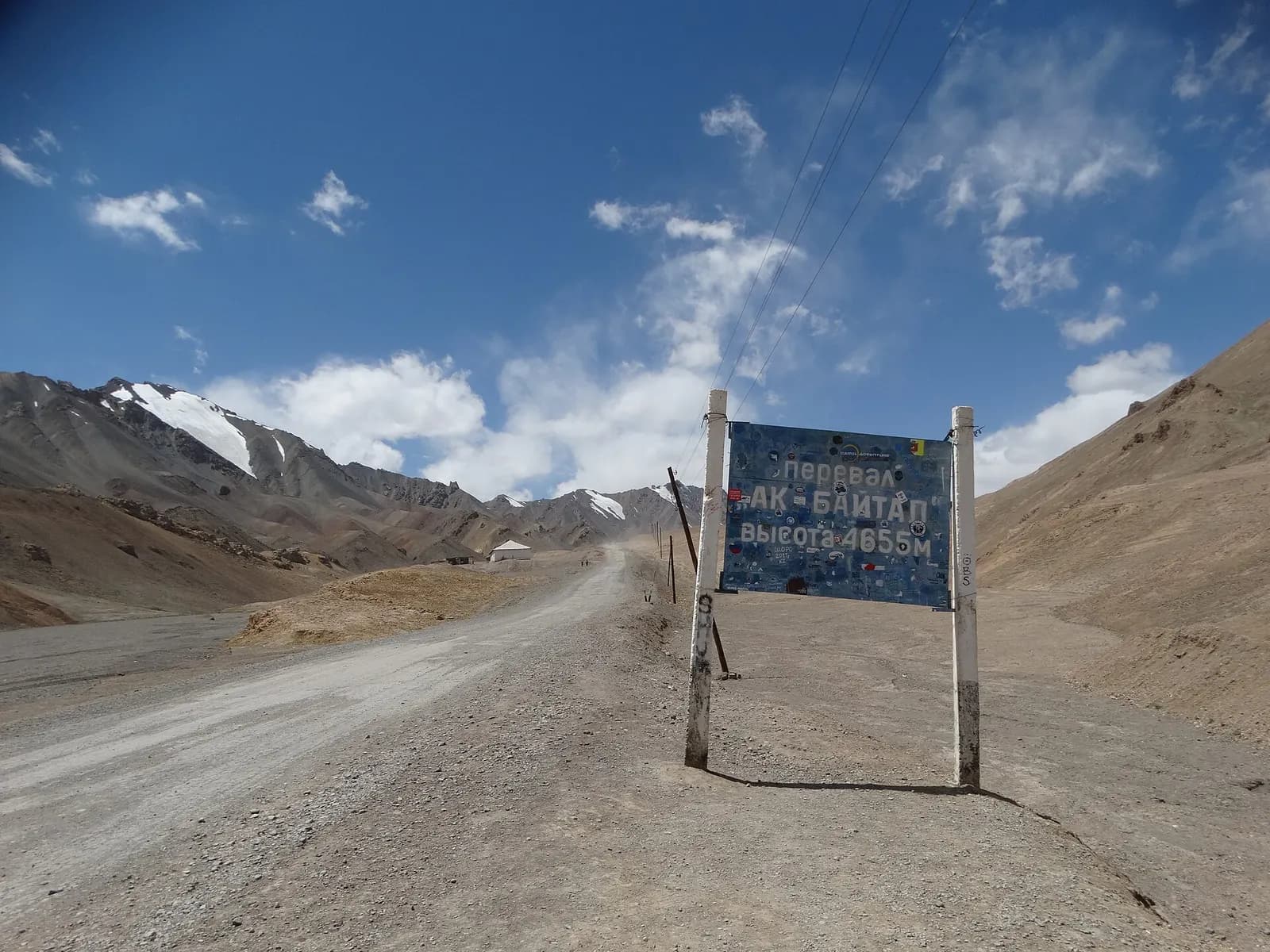 A gravel road and the sign marking the Ak-Baital Pass at 4,655 m on the Pamir Highway, with brown and snow-streaked mountains behind