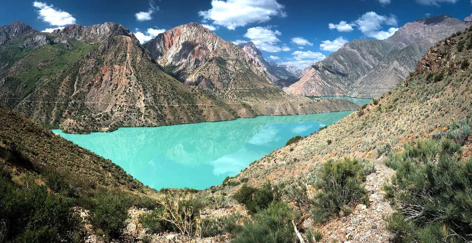 A turquoise lake filling a steep valley below red and grey rock peaks in the Fann Mountains, Tajikistan, with a hillside path in the foreground