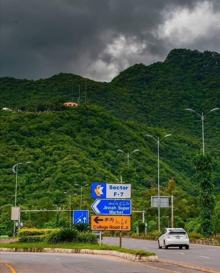 A road below the wooded Margalla Hills in Islamabad, sector signposts under a stormy sky