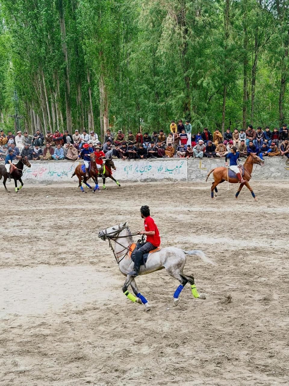 A village polo match in Khaplu, riders on the sand ground with spectators along the wall beneath poplars