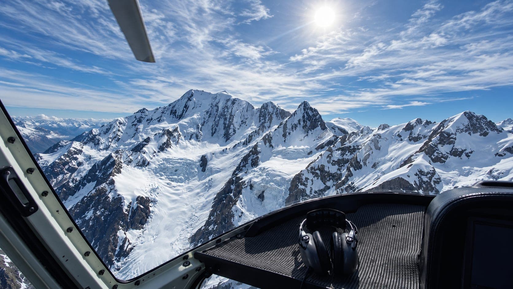 The view forward from a helicopter cockpit over snow-covered peaks and a glacier basin