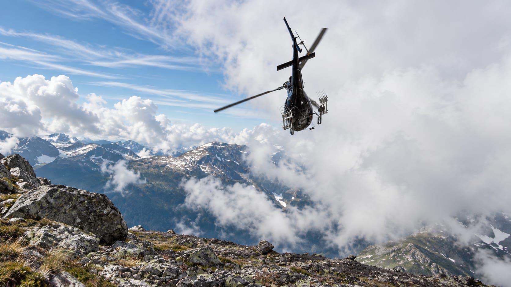 A helicopter lifting away from a rocky ridge with cloud and peaks beyond