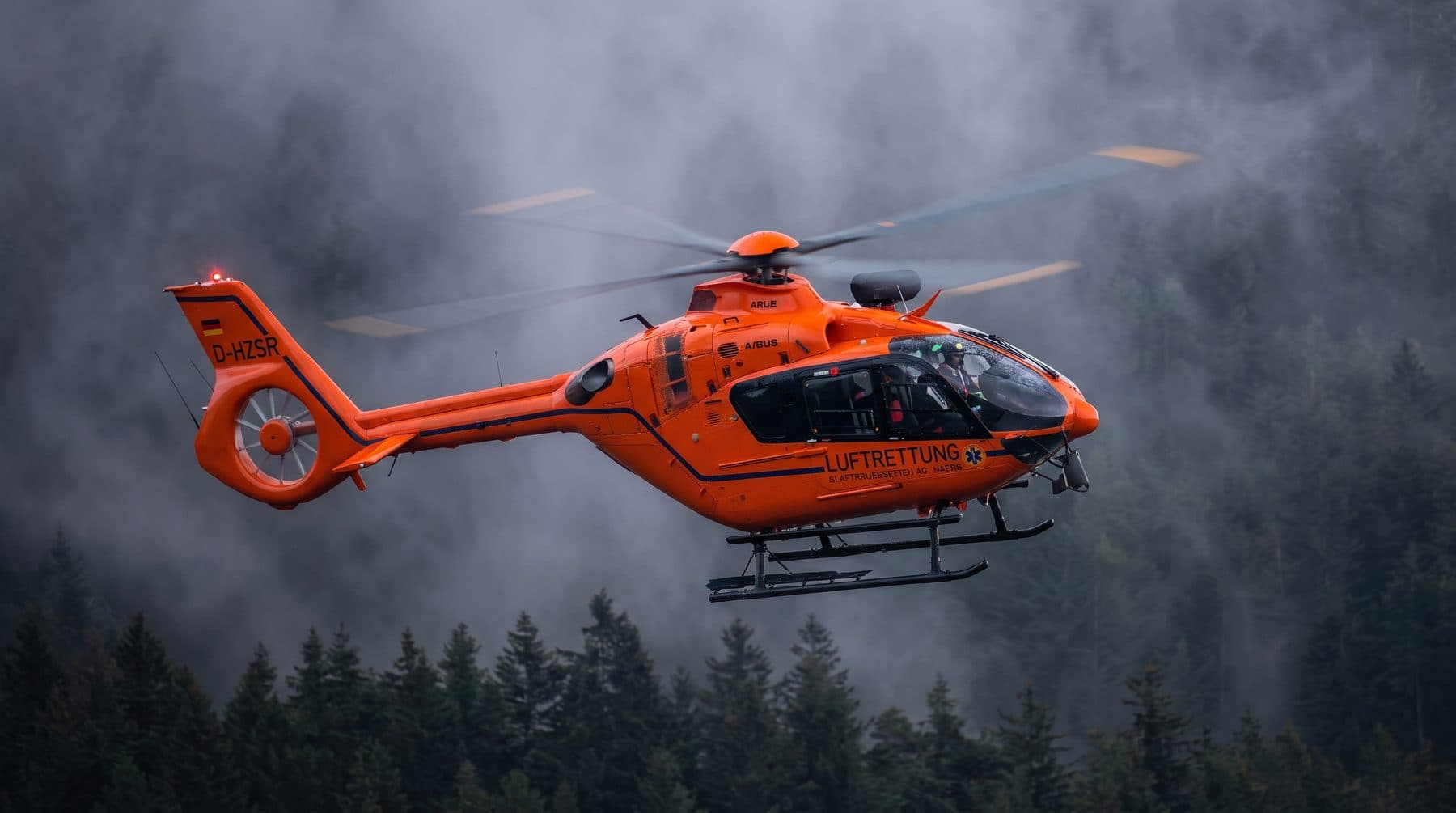 An orange helicopter flying through low cloud above a pine forest