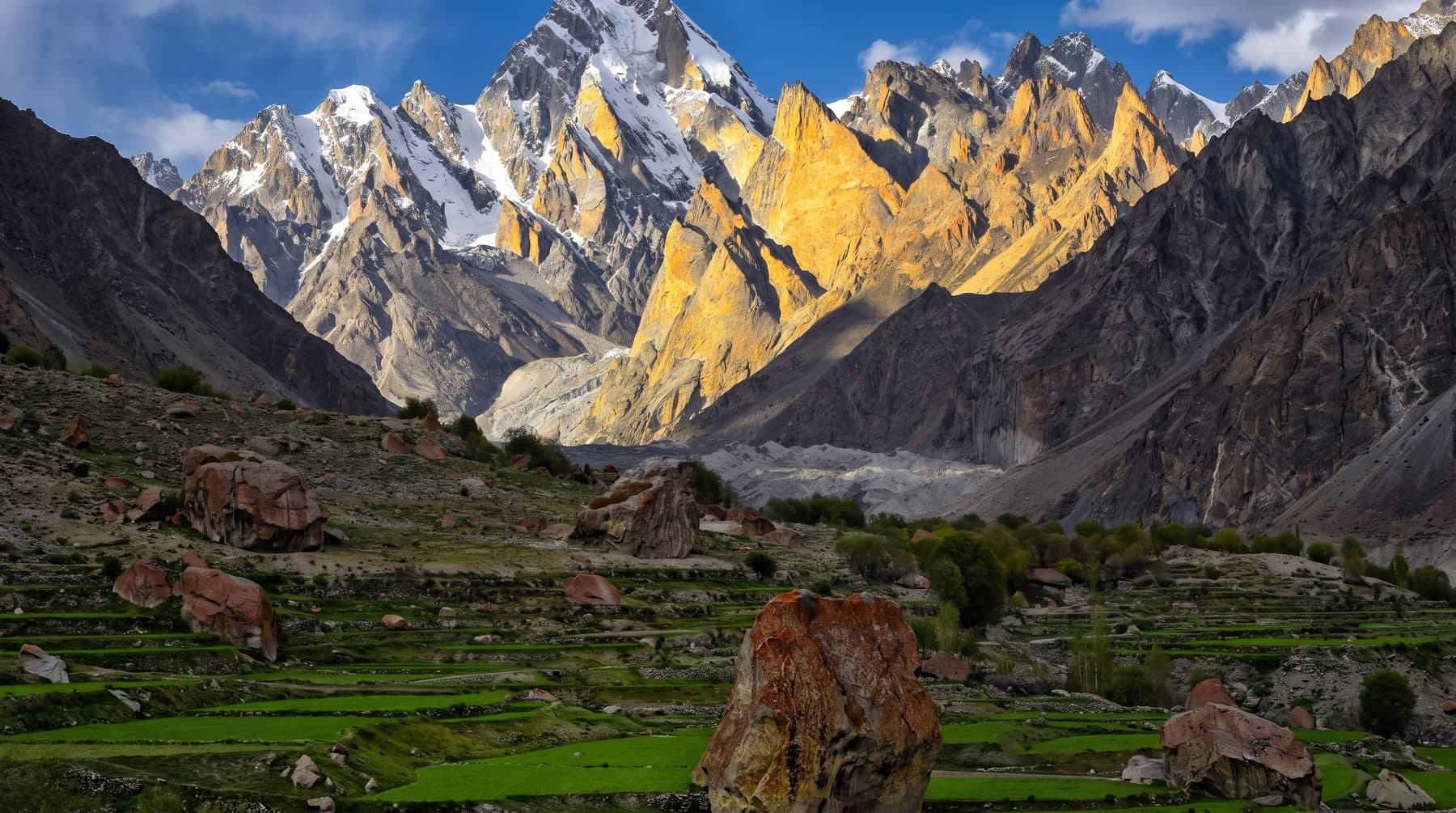 Golden granite spires above green terraced fields and boulders in a Karakoram valley