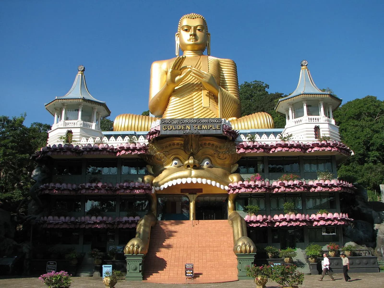 The modern Golden Temple building at the foot of the Dambulla cave temple, with a large gold seated Buddha on its roof under a blue sky