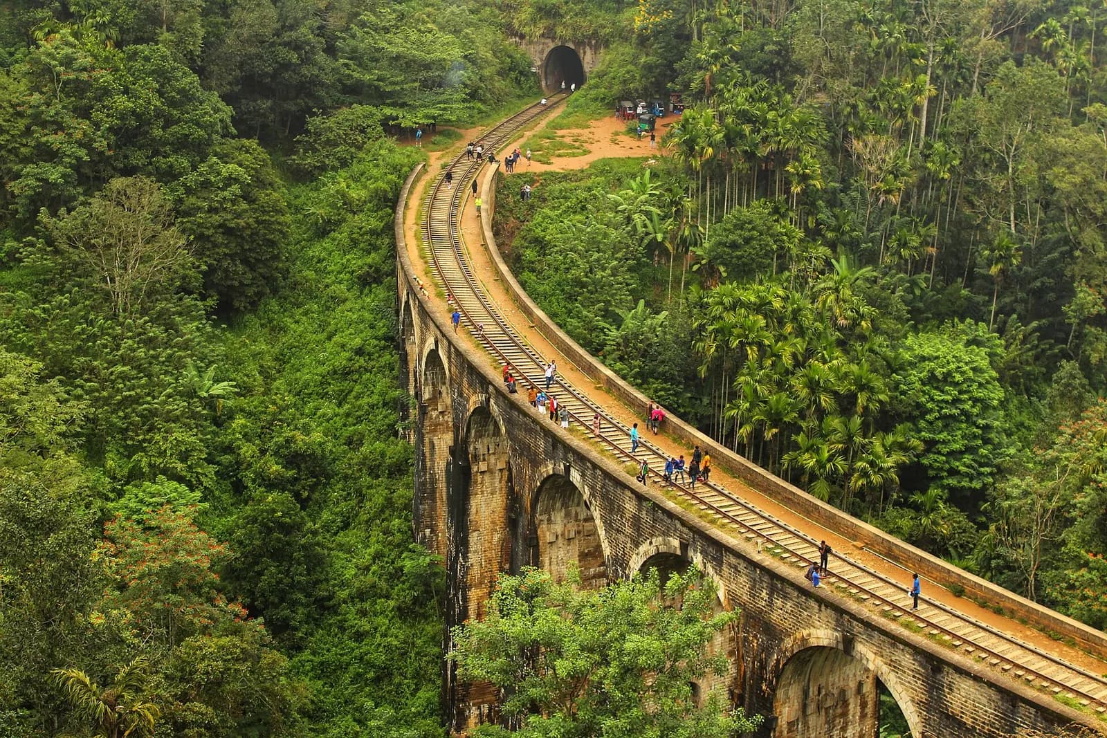The curved stone Nine Arch Bridge at Demodara near Ella, with people walking along the railway track between dense tropical forest and a tunnel mouth beyond