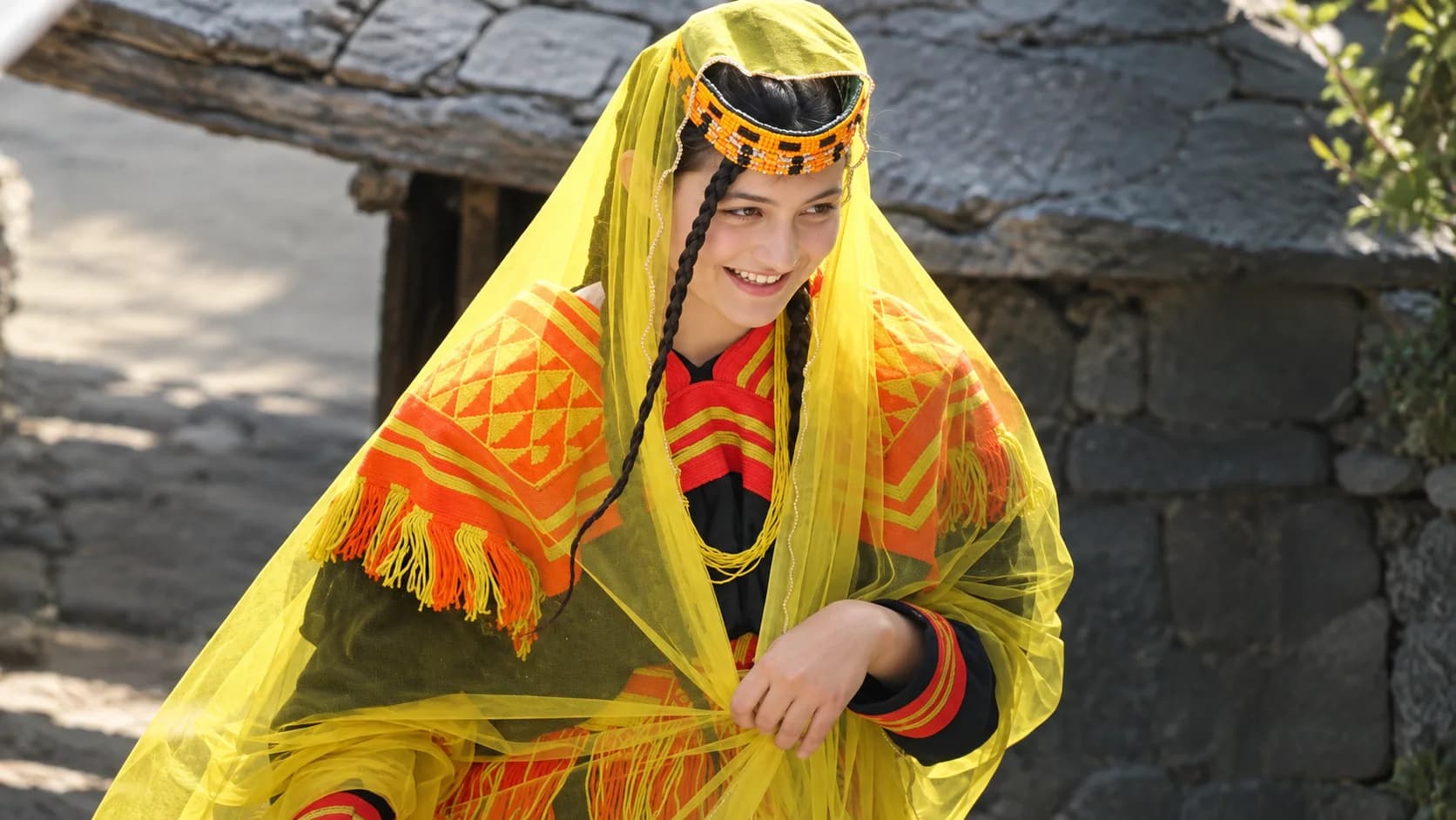 A smiling young Kalasha woman in Chitral wearing traditional black and bright orange embroidered clothing, covered with a sheer yellow veil and a beaded headdress.