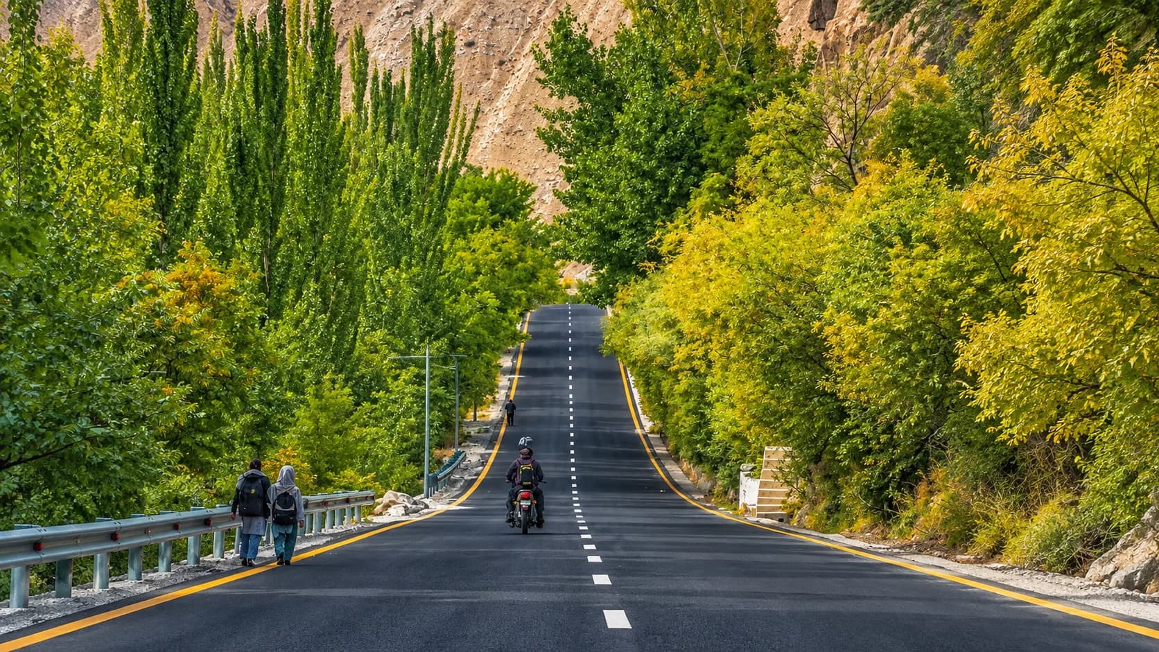 A motorcyclist and two pedestrians traveling up a beautifully paved section of a mountain road in northern Pakistan, surrounded by a lush green forest transitioning into early autumn colors beneath steep, rocky canyon walls.