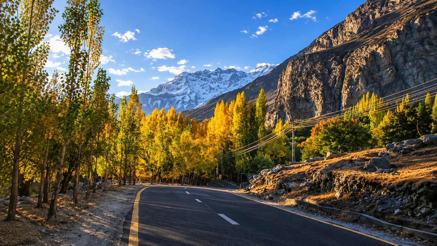 The Karakoram Highway winding through a mountain valley bathed in beautiful golden-hour sunlight, lined with vibrant yellow autumn trees and backed by a majestic snow-dusted mountain peak in Pakistan.