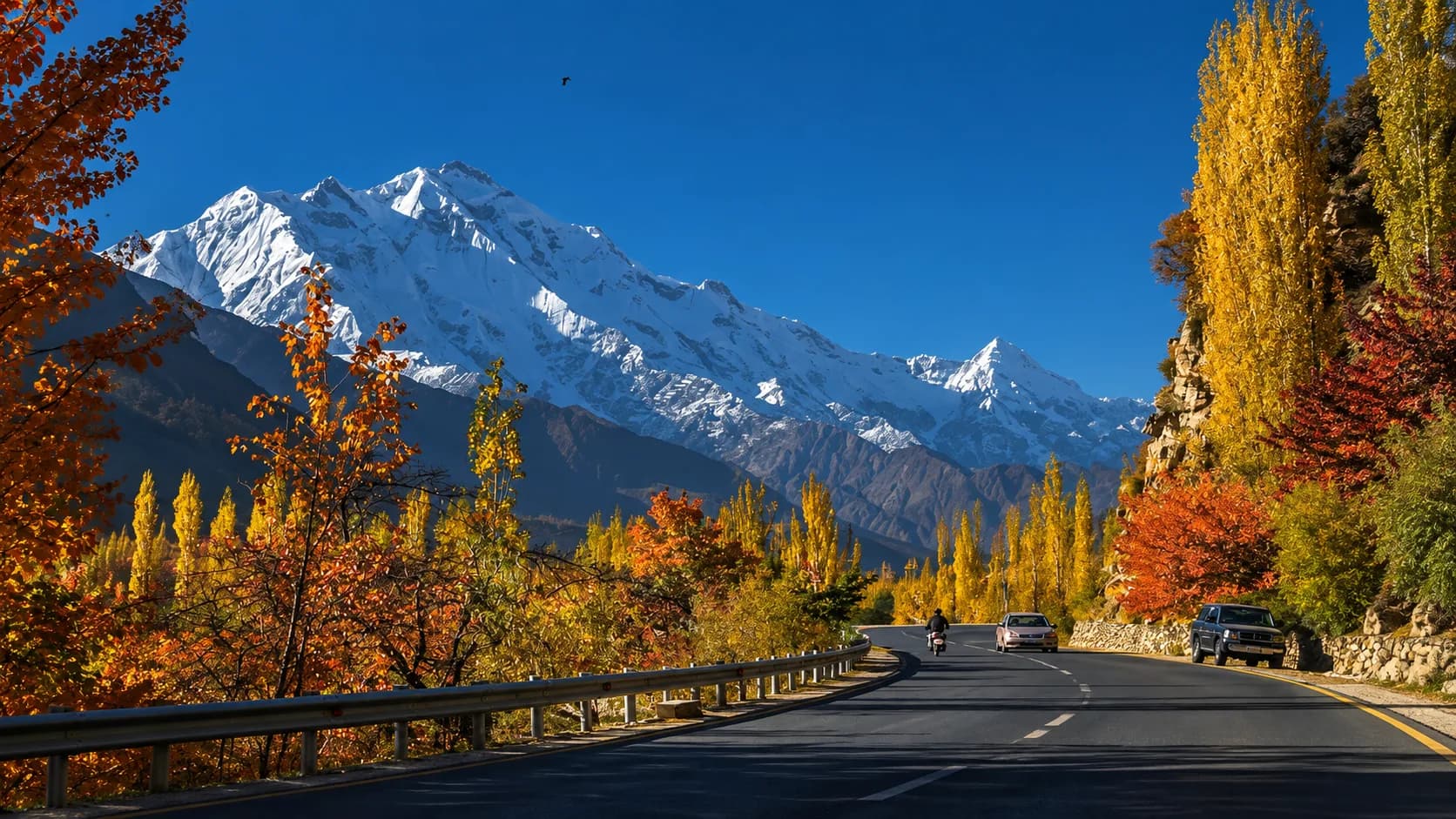 A scenic autumn view of the Karakoram Highway curving through Gilgit-Baltistan, featuring a car and a motorcycle driving past bright yellow and red trees, with a colossal, sunlit snow-capped mountain peak towering against a deep blue sky.