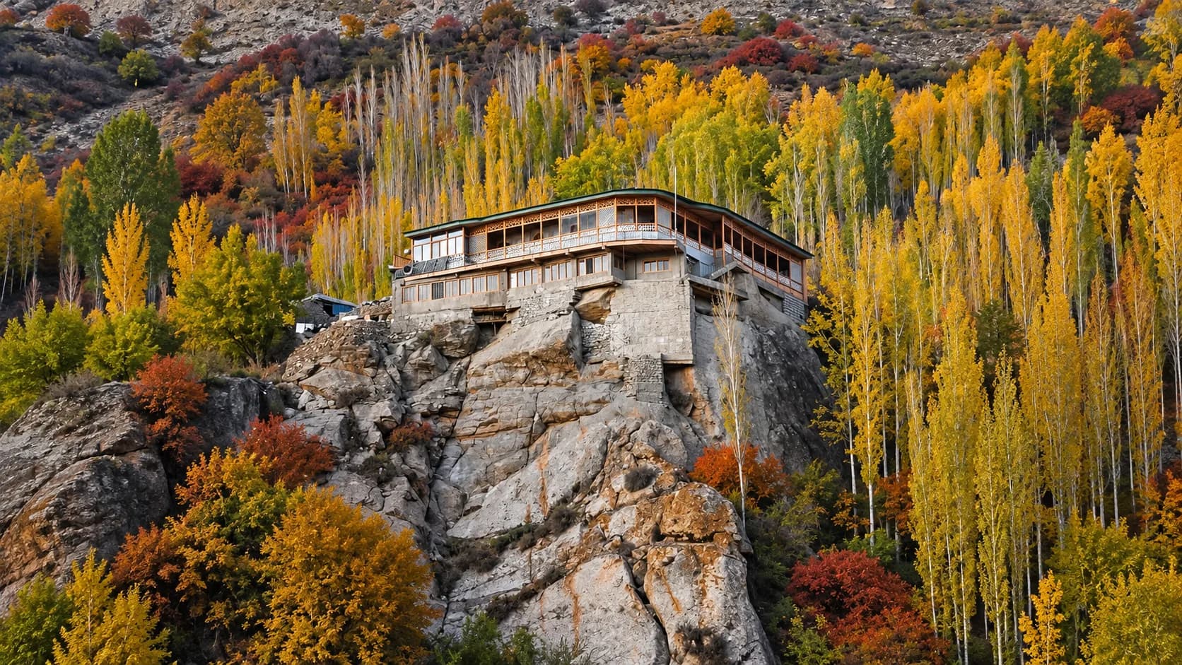 A traditional wooden and stone hotel building perched precariously on a steep, rocky cliffside, surrounded by vibrant yellow and orange autumn trees in the Hunza Valley.