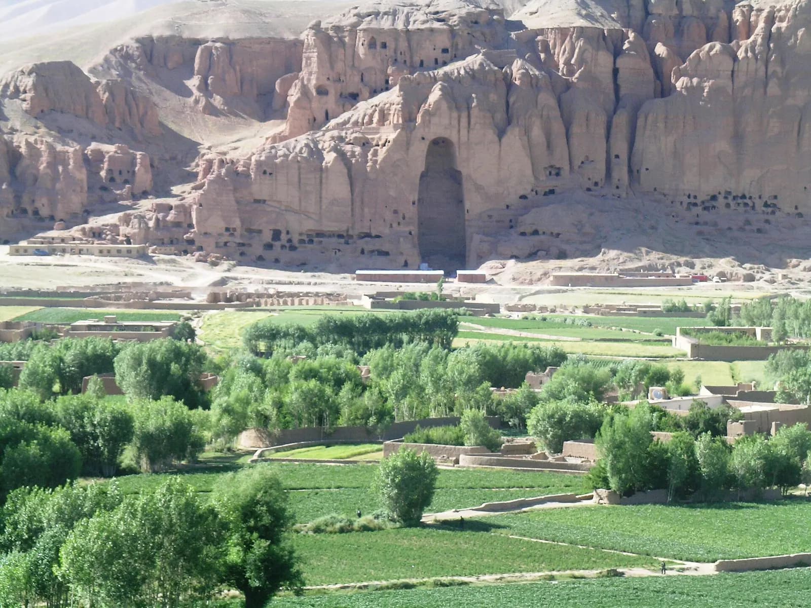 The empty niche of the larger Bamiyan Buddha in a sandstone cliff riddled with cave monasteries, above green fields and poplars in the Bamiyan valley