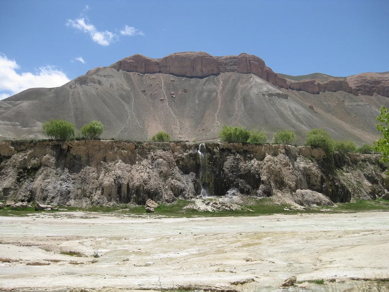 Water spilling over a natural travertine wall at Band-e-Amir, beneath a long ridge of red sandstone cliffs and a blue sky