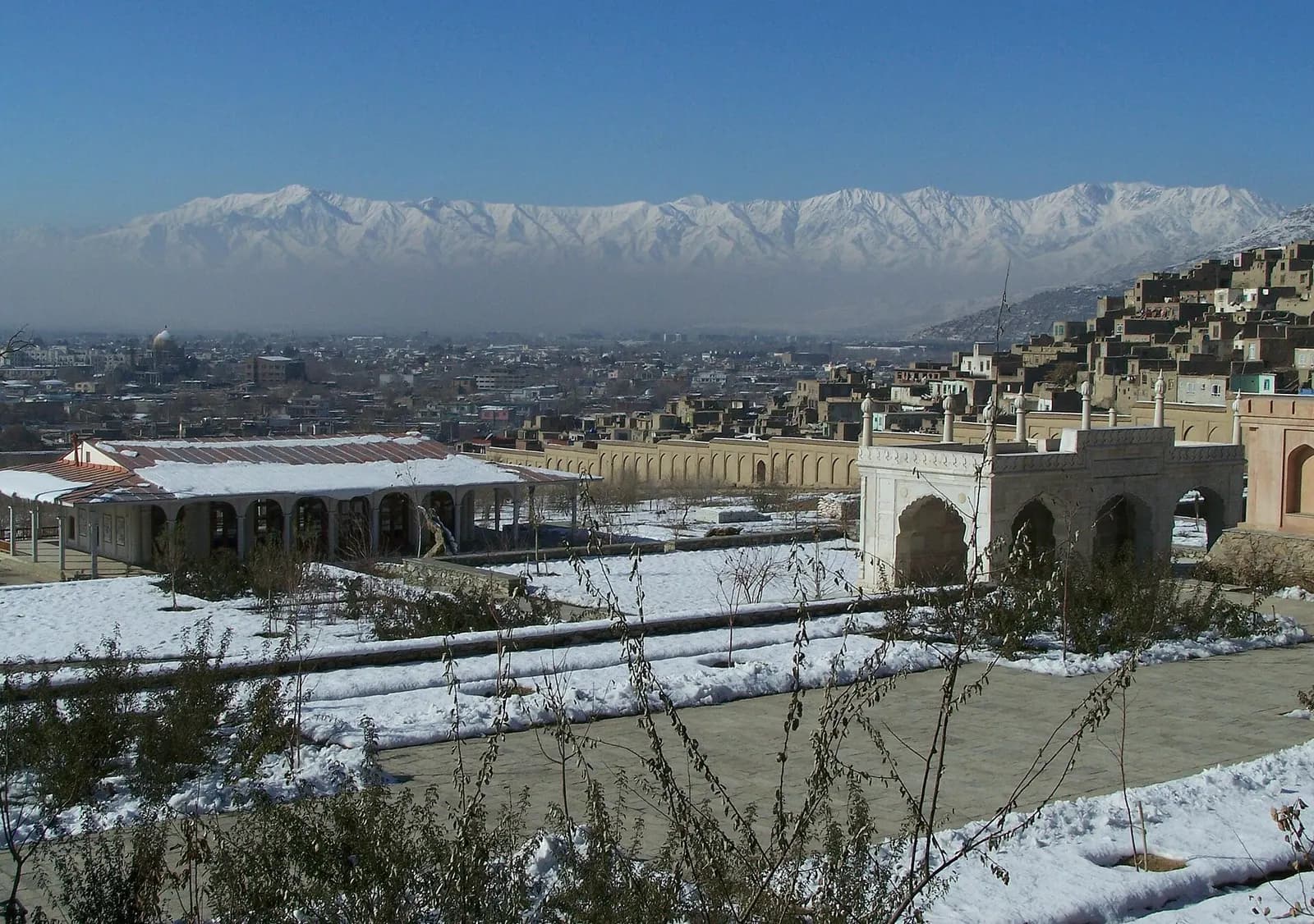 The terraced Gardens of Babur in Kabul under winter snow, with the white marble Shah Jahan mosque, the city beyond and a snow-covered range on the horizon
