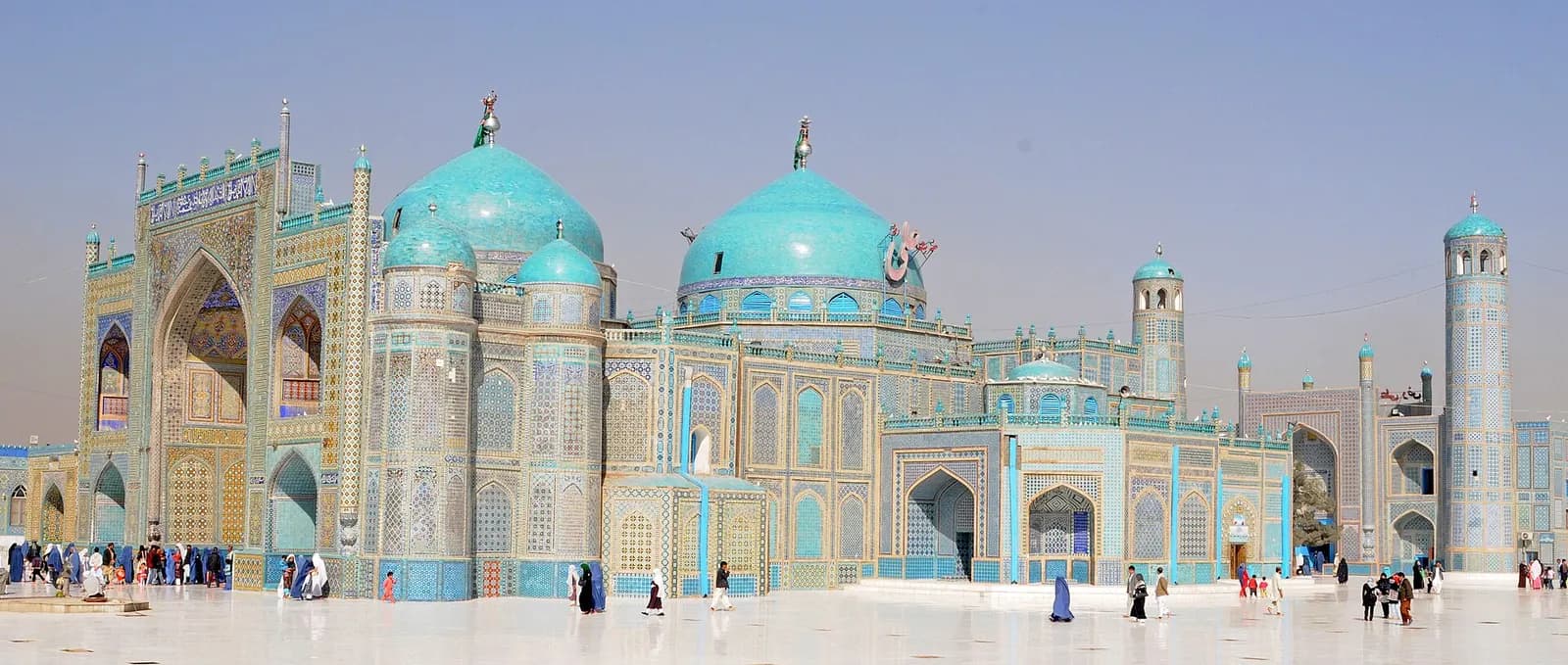 The turquoise domes and tiled facades of the Blue Mosque in Mazar-i-Sharif, with visitors crossing the white marble courtyard