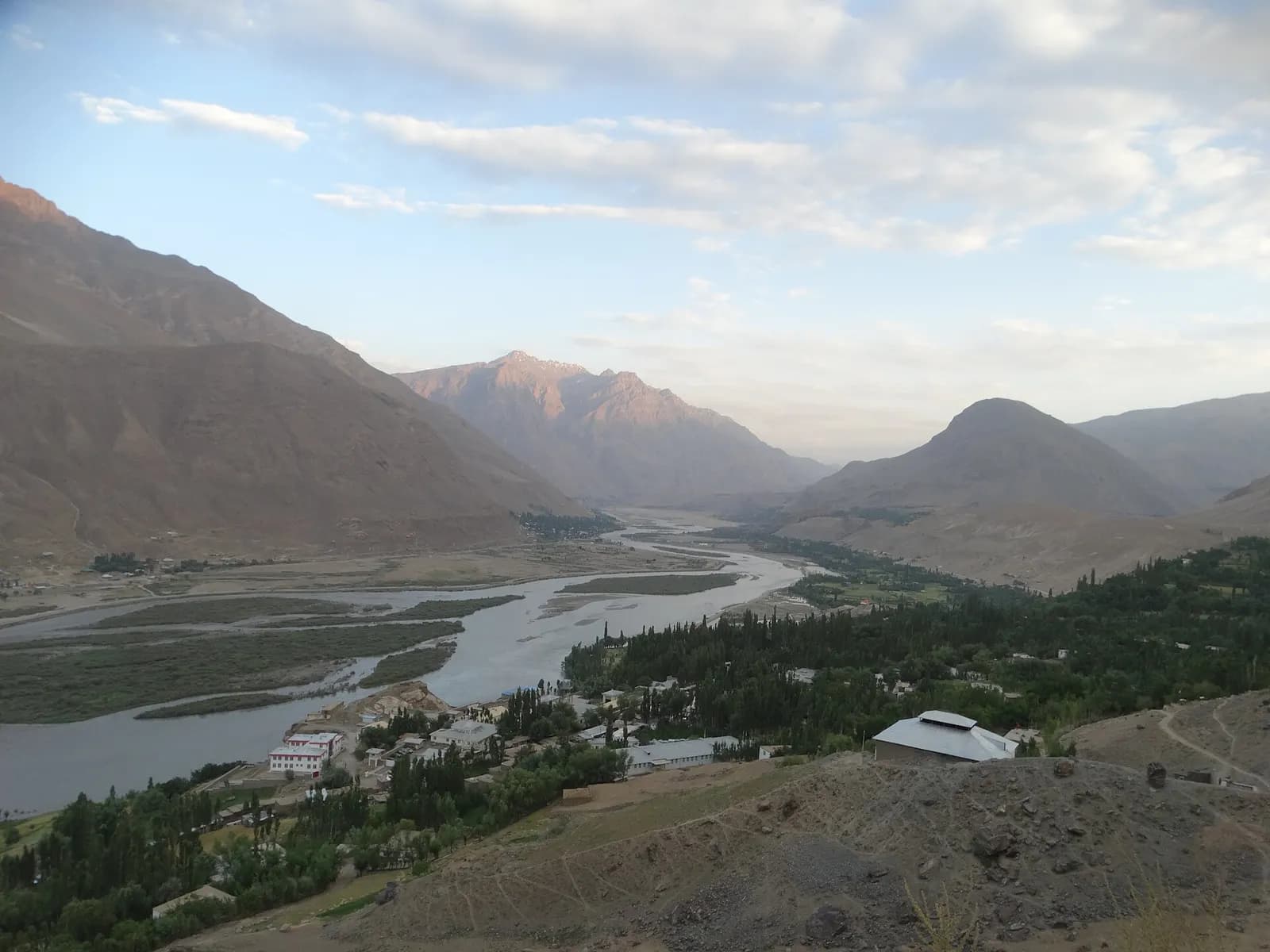 The braided Panj river winding between bare mountains near Qala-e Panja in the Afghan Wakhan, with a village of poplars and tin roofs on the near bank