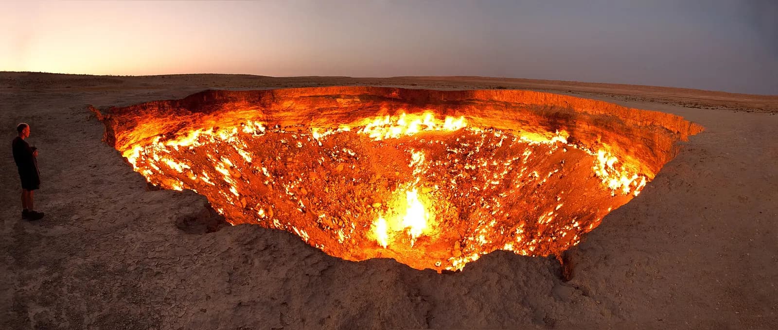 The burning Darvaza gas crater at dusk in the flat Karakum desert, Turkmenistan, with a lone figure standing at the rim