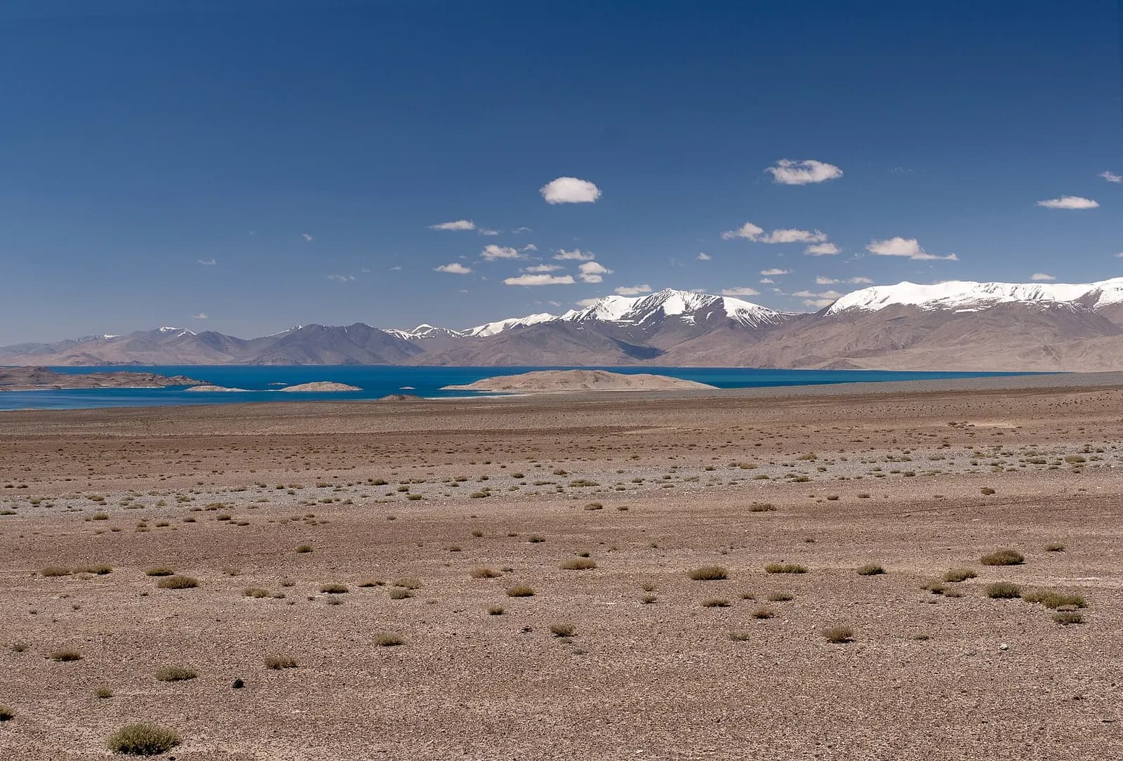 Karakul lake, deep blue below snow-capped ridges, seen across the stony high plateau beside the Pamir Highway in Tajikistan