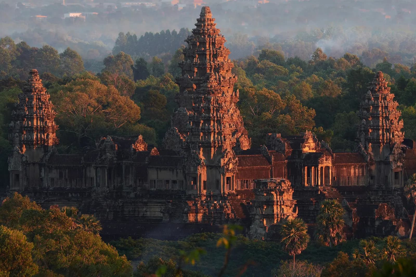 The towers of Angkor Wat lit orange by low sun above the surrounding forest, seen from Phnom Bakheng hill, Cambodia