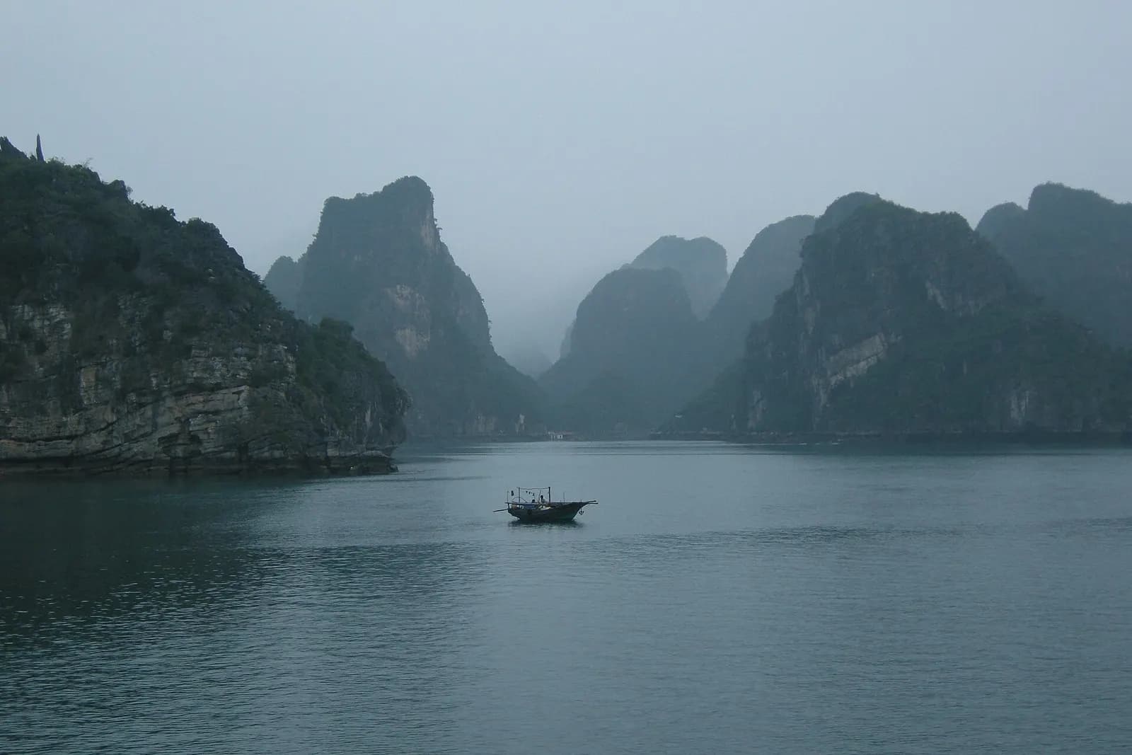 A small fishing boat on still water between steep limestone islands in morning mist, Ha Long Bay, Vietnam