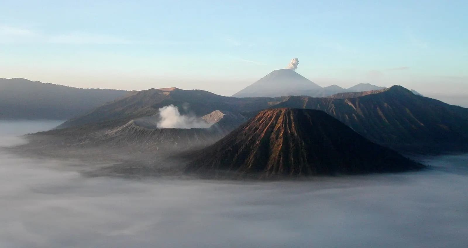 Mount Bromo's smoking crater and the ribbed cone of Mount Batok rising from mist in the Tengger caldera at sunrise, with Mount Semeru venting in the distance, East Java