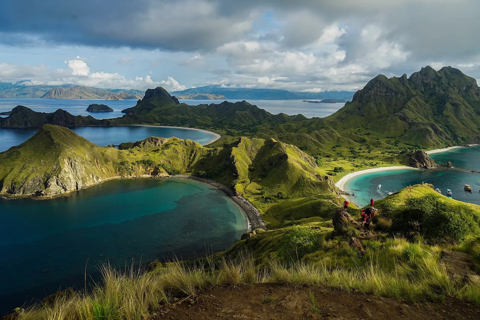 Grassy ridges and curved beaches around the bays of Padar Island, with boats anchored offshore and walkers on the viewpoint trail, Komodo National Park