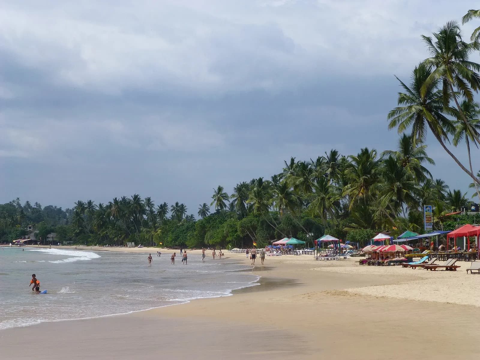 The curve of Mirissa beach with coconut palms, beach umbrellas and people wading in the surf under a cloudy sky