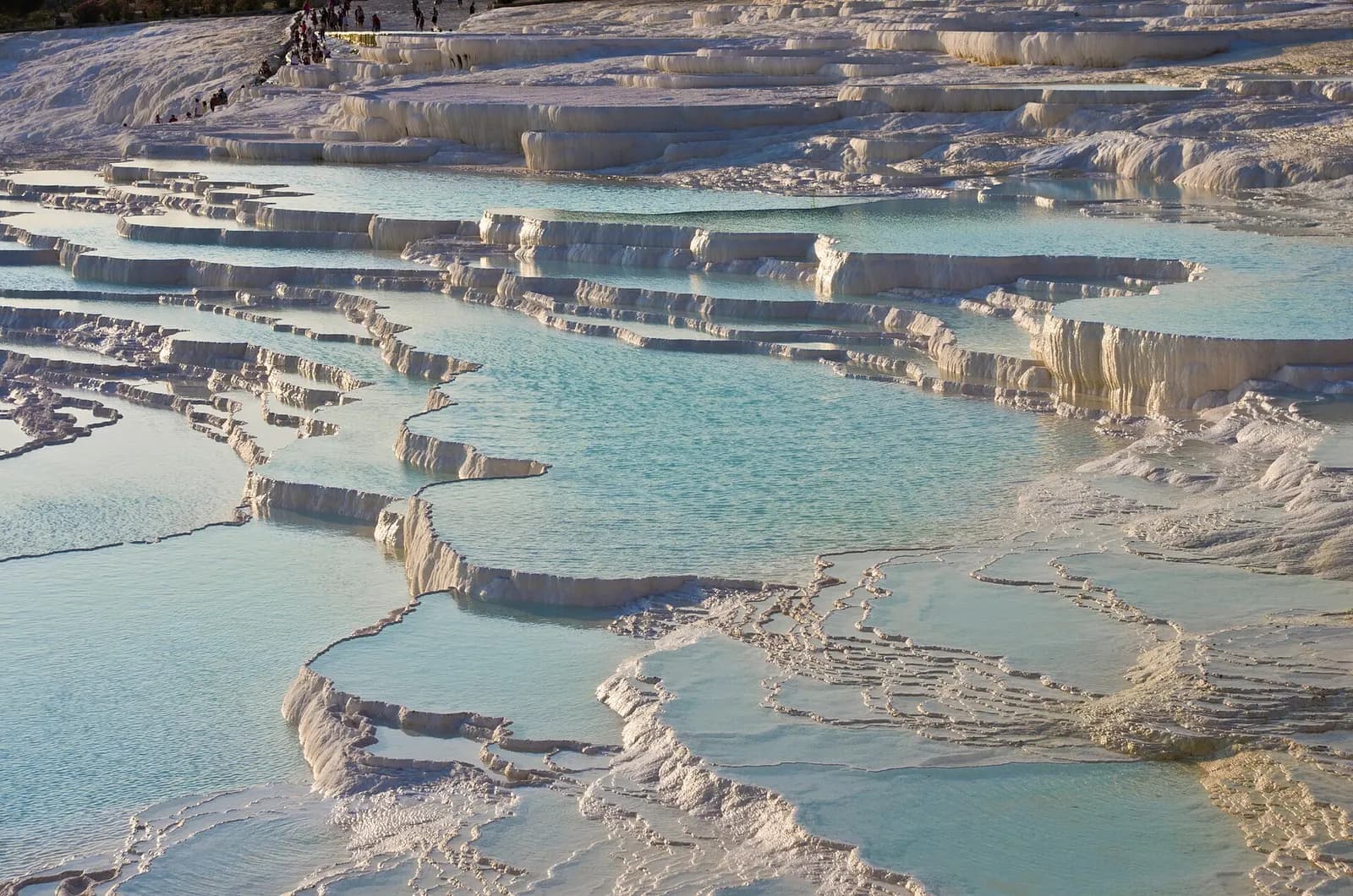Tiers of white travertine terraces filled with pale blue water at Pamukkale, with visitors walking along the top