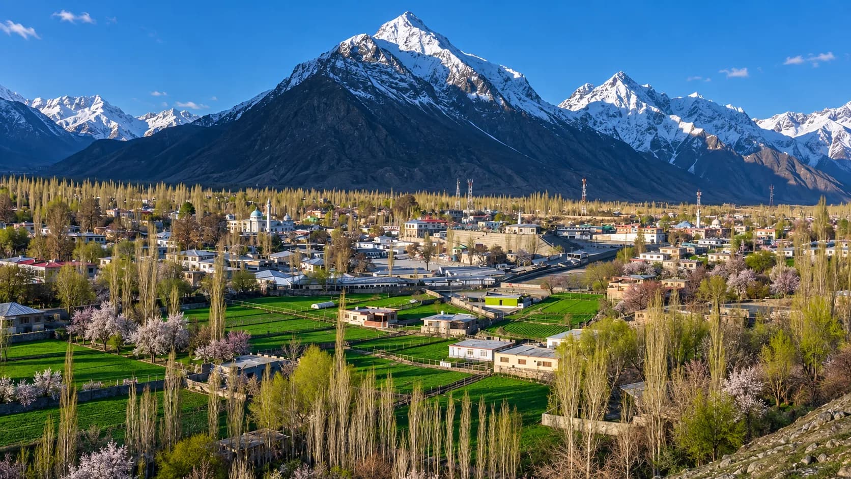 High-angle perspective of a pastoral mountain settlement in northern Pakistan with patchwork green fields and blooming orchards, set beneath a sharp, snow-capped mountain ridge.