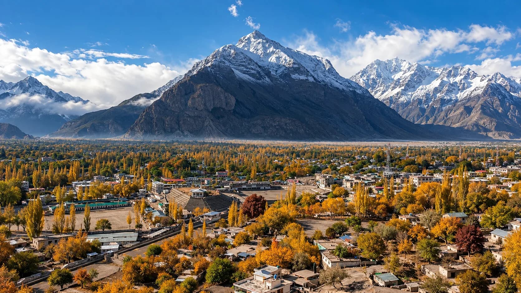 An elevated panoramic view over Skardu town ablaze with golden-yellow autumn poplar trees and terraced fields, framed by a prominent snow-draped pyramid mountain under a blue sky.
