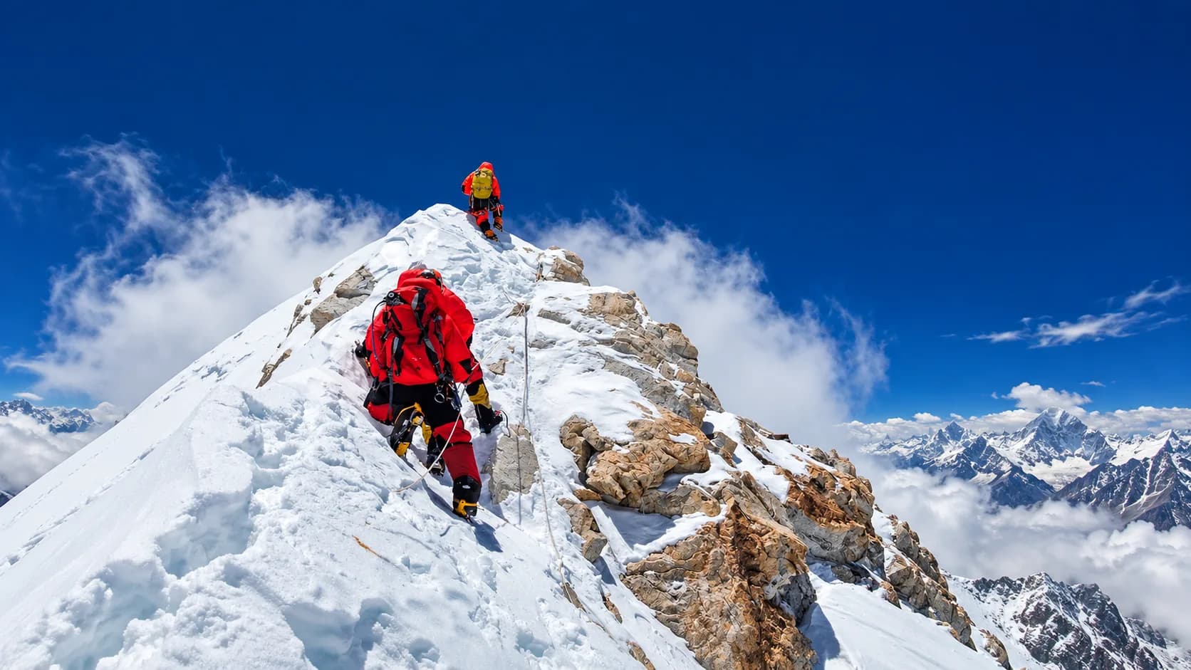 Climbers wearing red and yellow insulated suits navigating a knife-edge snow and rock summit ridge clipped to fixed ropes, surrounded by clouds and distant mountain summits under a clear blue sky.