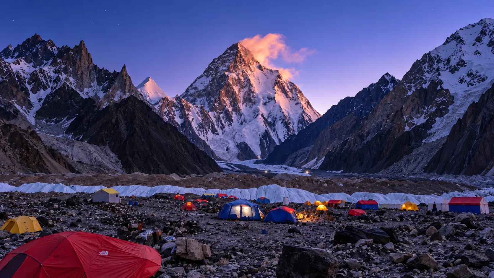 Illuminated expedition dome and mess tents glowing at dusk across the rocky glacial moraine of K2 Base Camp, with soft pink alpenglow lighting the snow-covered summit of K2 against a deep twilight sky.